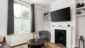 A living room with a fireplace, a TV mounted on the wall, a round coffee table, and a grey armchair. - New! Modern 2BR House w/ Patio in Acton (Greater London)