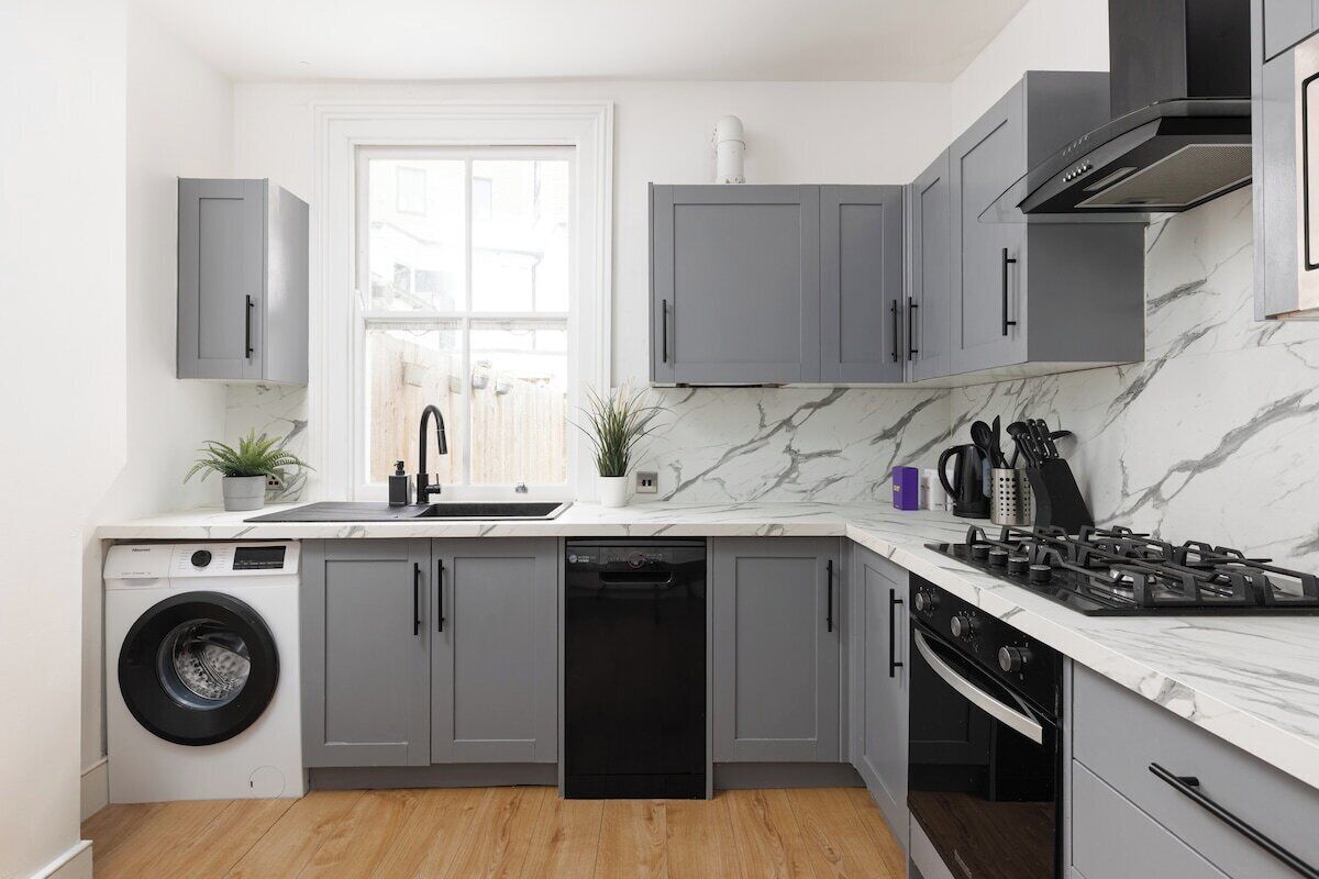 Une cuisine compacte avec des meubles gris, un lave-linge encastré, un lave-vaisselle, une cuisinière à gaz et une fenêtre qui laisse entrer la lumière naturelle.
