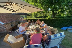 Eine Gruppe von Personen speist draußen an einem Tisch unter einem großen Sonnenschirm, umgeben von Korbstühlen und einem Backsteinhaus.