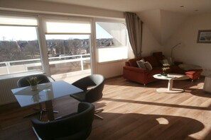 A modern living room with a dining table, two black chairs, a red sofa, a small white coffee table, and large windows with a view of buildings and trees. - 347 in the Trafalgar House (Cuxhaven - Döse)