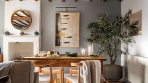 A dining area with a wooden table, chairs, a fireplace, a large painting, and a potted tree.
