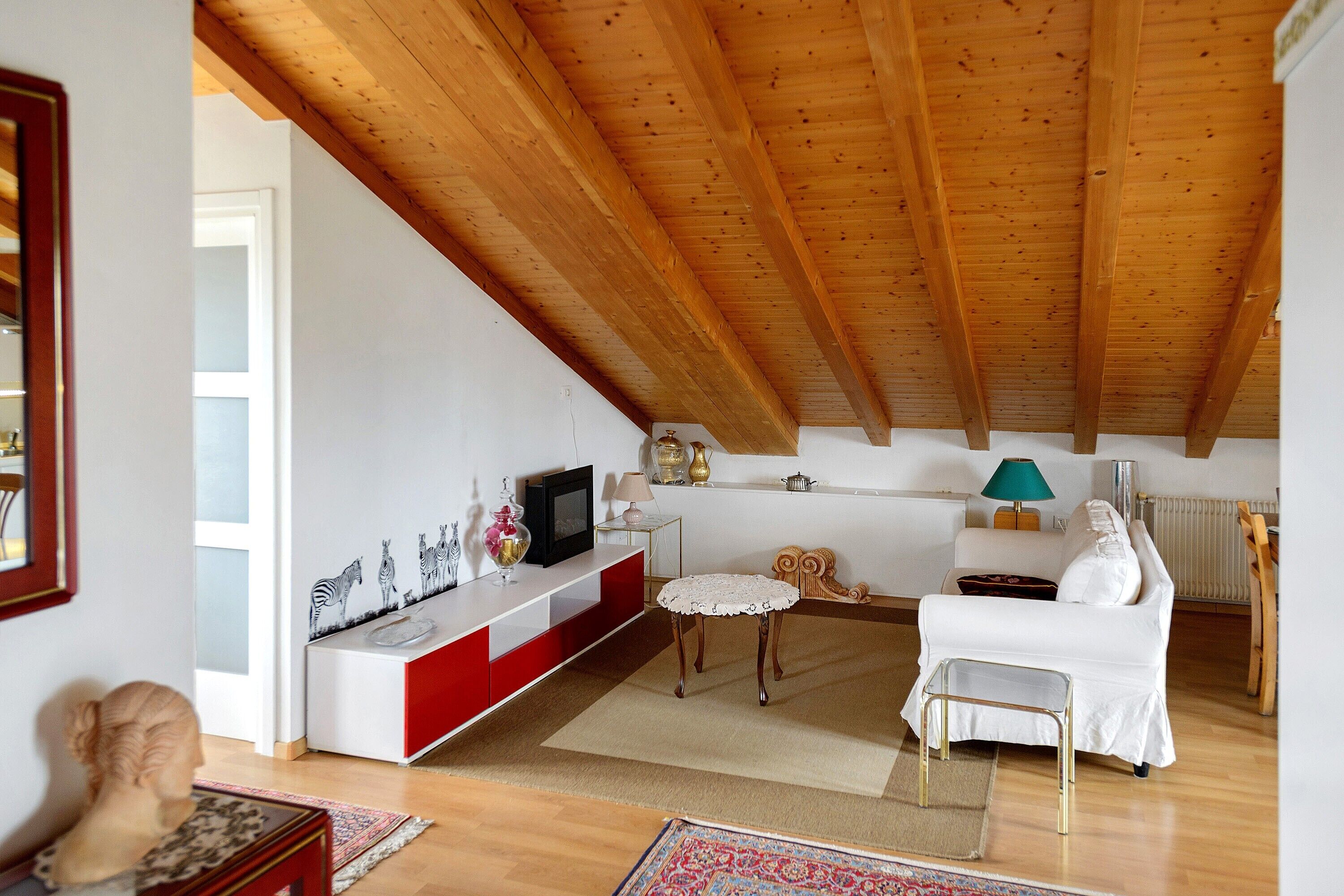 Un salon avec un plafond en bois, un canapé blanc, une console rouge et blanche, et une petite table avec une lampe.