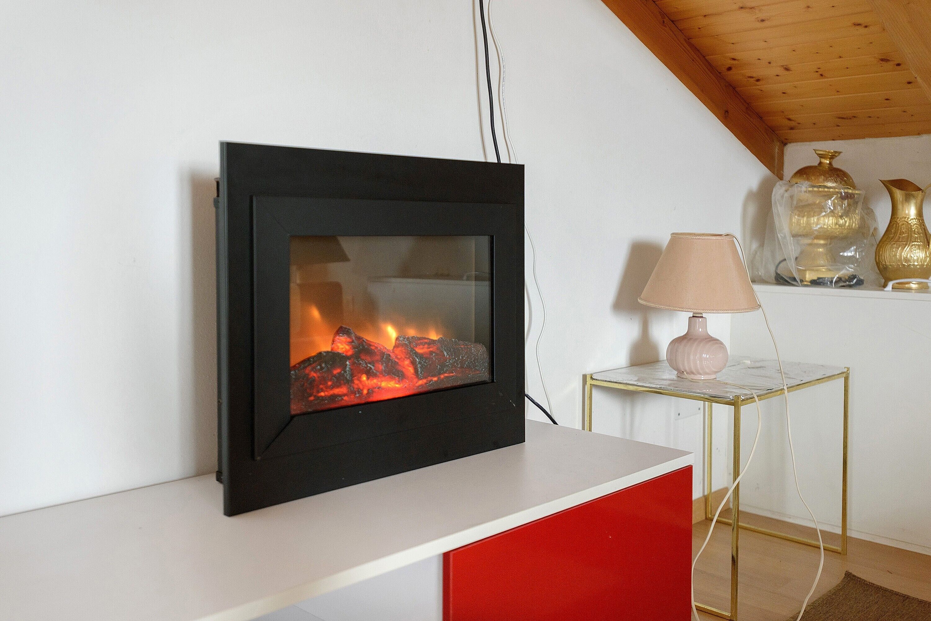 Een open haard met vlammen, een witte muur, een rode kast, een tafel met gouden lijst en een lamp, en decoratieve vazen.