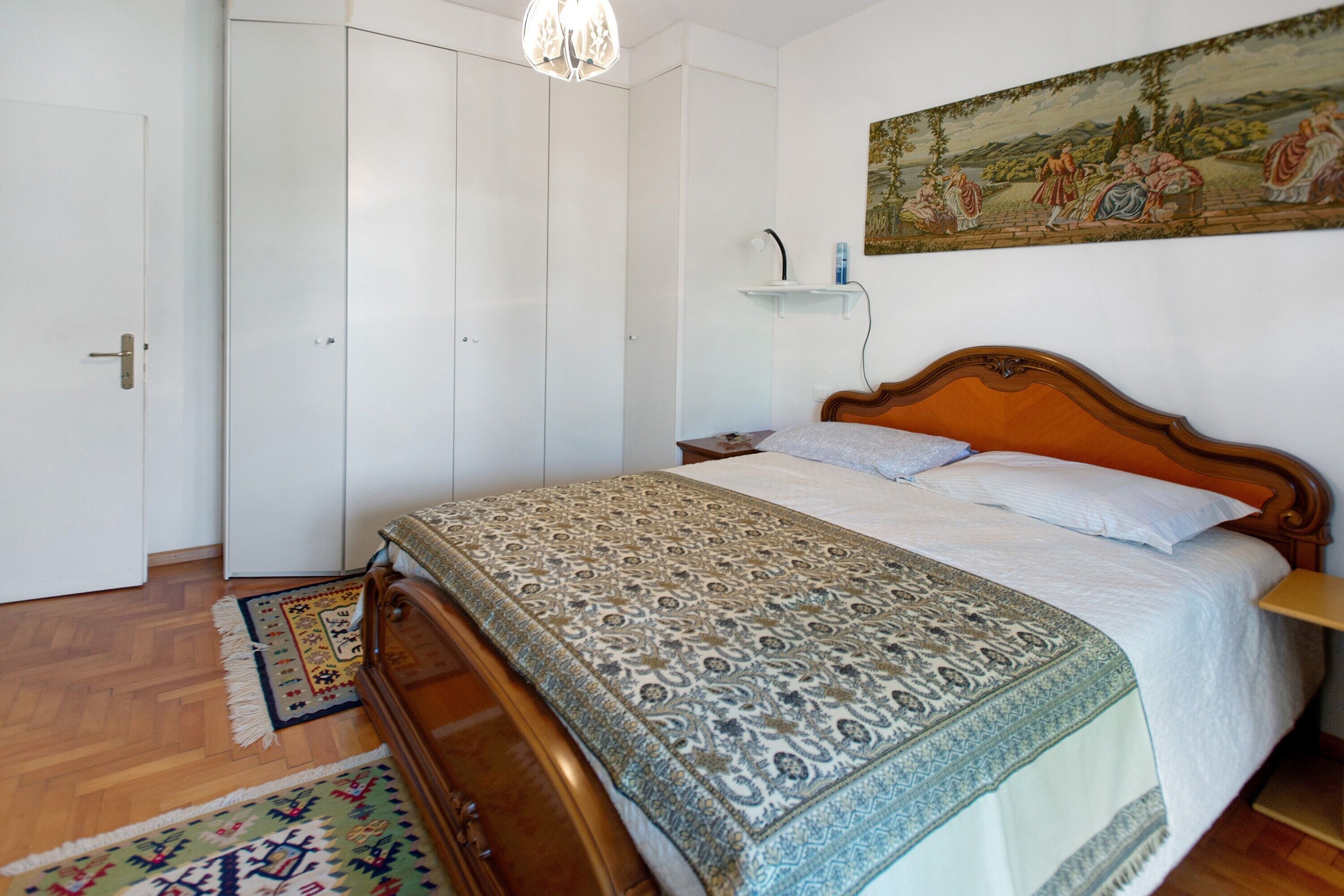 Ein Schlafzimmer mit einem hölzernen Bett, einem Nachttisch, einem an der Wand befestigten Regal, einer Tür und einem gemusterten Teppich.