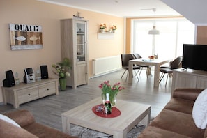 Ein Wohnzimmer mit einer Couch, einem Couchtisch mit einer Flasche, einem Schrank, einem Fernseher und einem Essbereich mit Stühlen und einem Tisch.