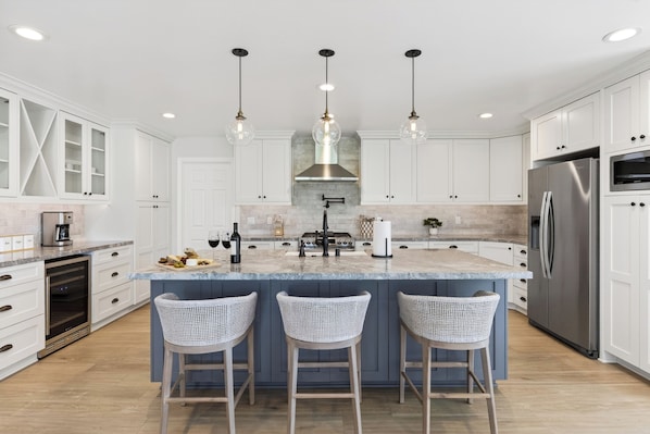 Una cocina moderna con armarios blancos, una isla central con taburetes de bar, un refrigerador de acero inoxidable e iluminación colgante.