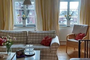 Ein Wohnzimmer mit einer Couch, einem Sessel, einem Couchtisch und zwei Fenstern mit Vorhängen.