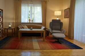Ein Wohnzimmer mit Sofa, Sessel, Tisch, Lampe, Bücherregal und Blick durch das Fenster.