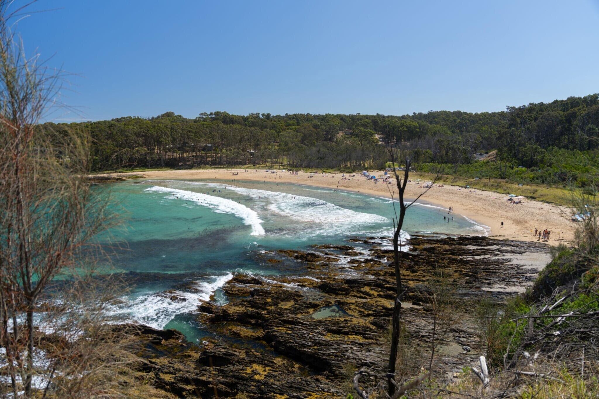 A coastal beach with clear turquoise waters, rocky shoreline, and a forested hill in the background.
