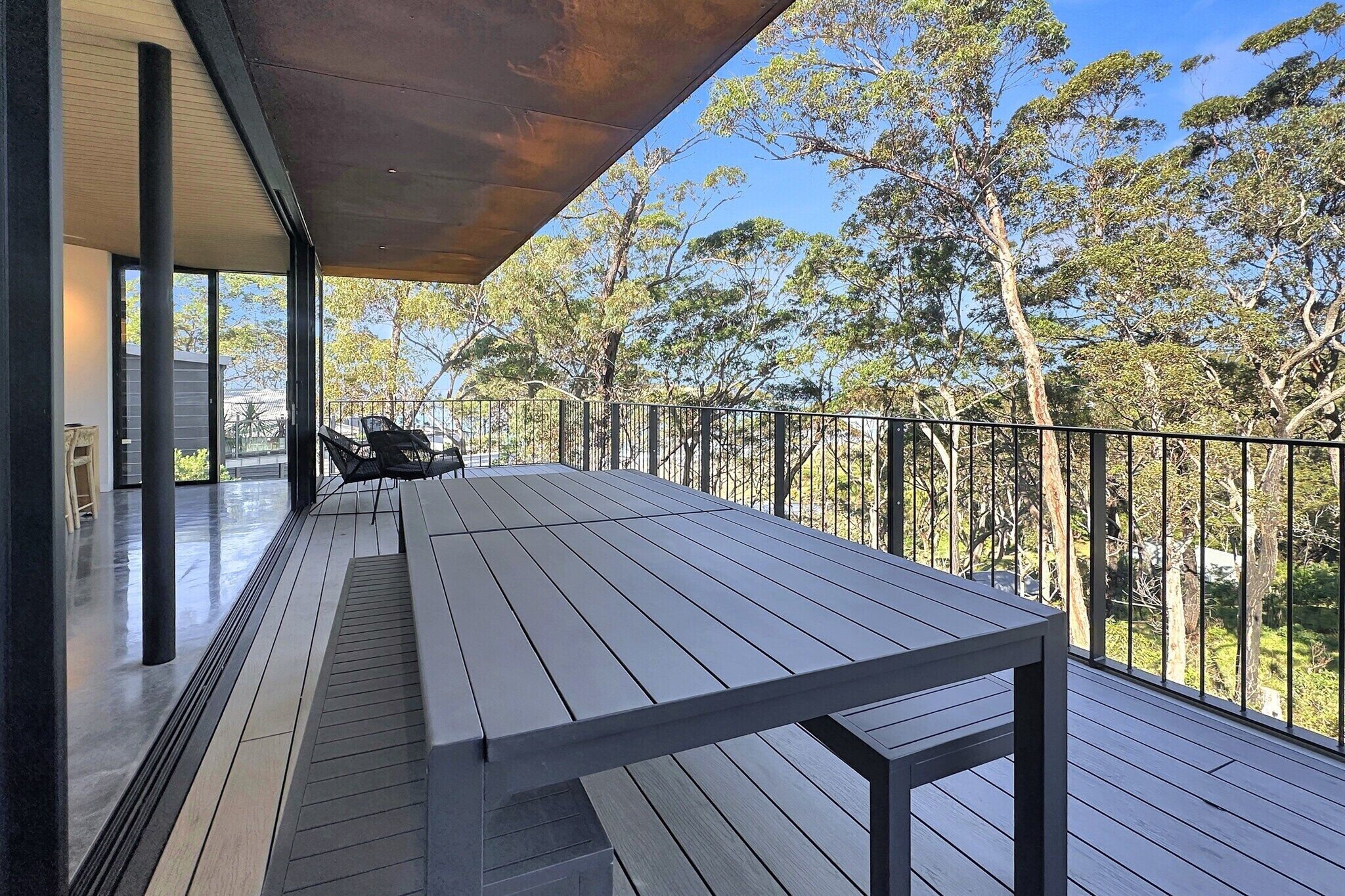 A wooden deck with a table and chairs, a metal railing, and a view of trees.