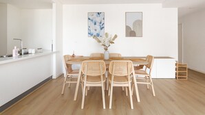 Un comedor moderno con una mesa blanca, seis sillas de mimbre, un jarrón con flores y dos cuadros en la pared.