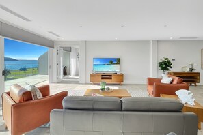 A modern living room with a large TV, leather sofas, a wooden coffee table, and a view of the ocean. - Coastal 3-Bed Retreat Near Batemans Bay (Batemans Bay)