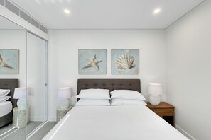 A bedroom with a bed, two bedside tables with lamps, a nightstand, a mirror, and two wall-mounted paintings of a starfish and a shell. - Coastal 3-Bed Retreat Near Batemans Bay (Batemans Bay)