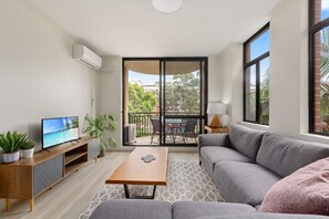 Una sala de estar moderna con un sofá gris, una mesa de centro de madera, un televisor sobre un mueble de madera, un balcón con mesa y sillas, y vistas a zonas verdes y edificios.