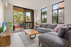 Un salón con un sofá gris, una mesa de centro de madera, un televisor sobre un mueble de madera, una lámpara y vistas a un balcón con mesa y sillas.
