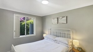 Ein Schlafzimmer mit einem Bett, einem Nachttisch, einer Lampe, einem Fenster mit Vorhängen und zwei an der Wand befestigten Bildern.