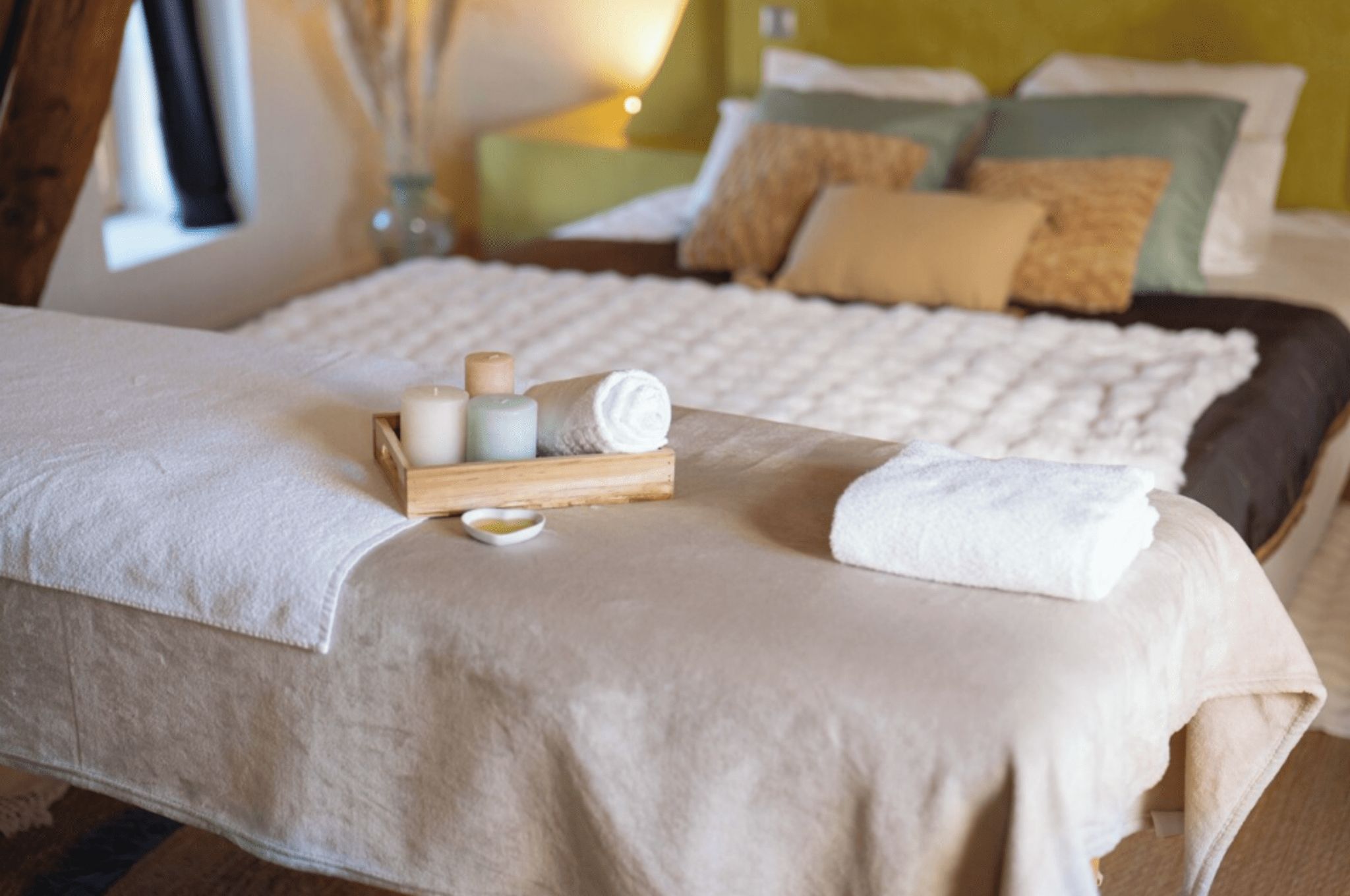 Een bed met een witte spreidek, een houten dienblad met kaarsen en een opgerolde handdoek.
