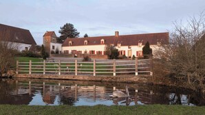 A traditional building with a red-tiled roof, a white facade, and a small tower. In front, there is a wooden bridge over a calm water body with a reflection of the building. - L'Atelier Vacation Rental (Wierre-Effroy)