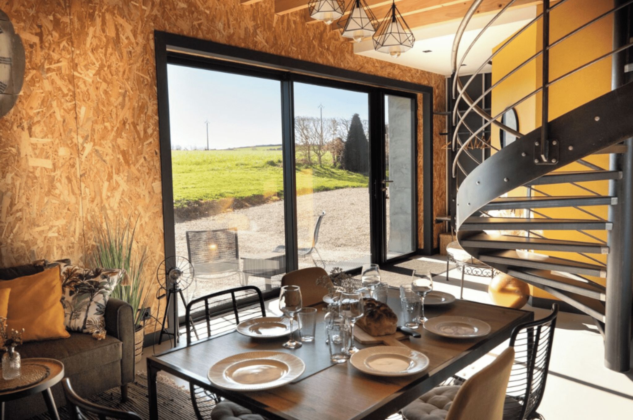 Een eetgedeelte met een tafel voor vier personen, een spiraaltrap en een groot raam met uitzicht op een grasveld.