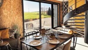 A dining area with a table set for four, a spiral staircase, and a large window with a view of a grassy field. - L'Atelier Vacation Rental (Wierre-Effroy)