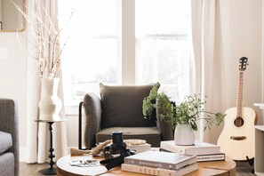 Ein gemütliches Wohnzimmer mit einem braunen Ledersessel, einem Couchtisch mit Büchern und einer Kamera, einer Gitarre und einer Pflanzvase.