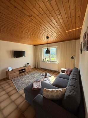 Ett vardagsrum med ett träpanelat tak, ett stort fönster med gardiner, en platt-TV, en soffa, ett soffbord och ett TV-bord.
