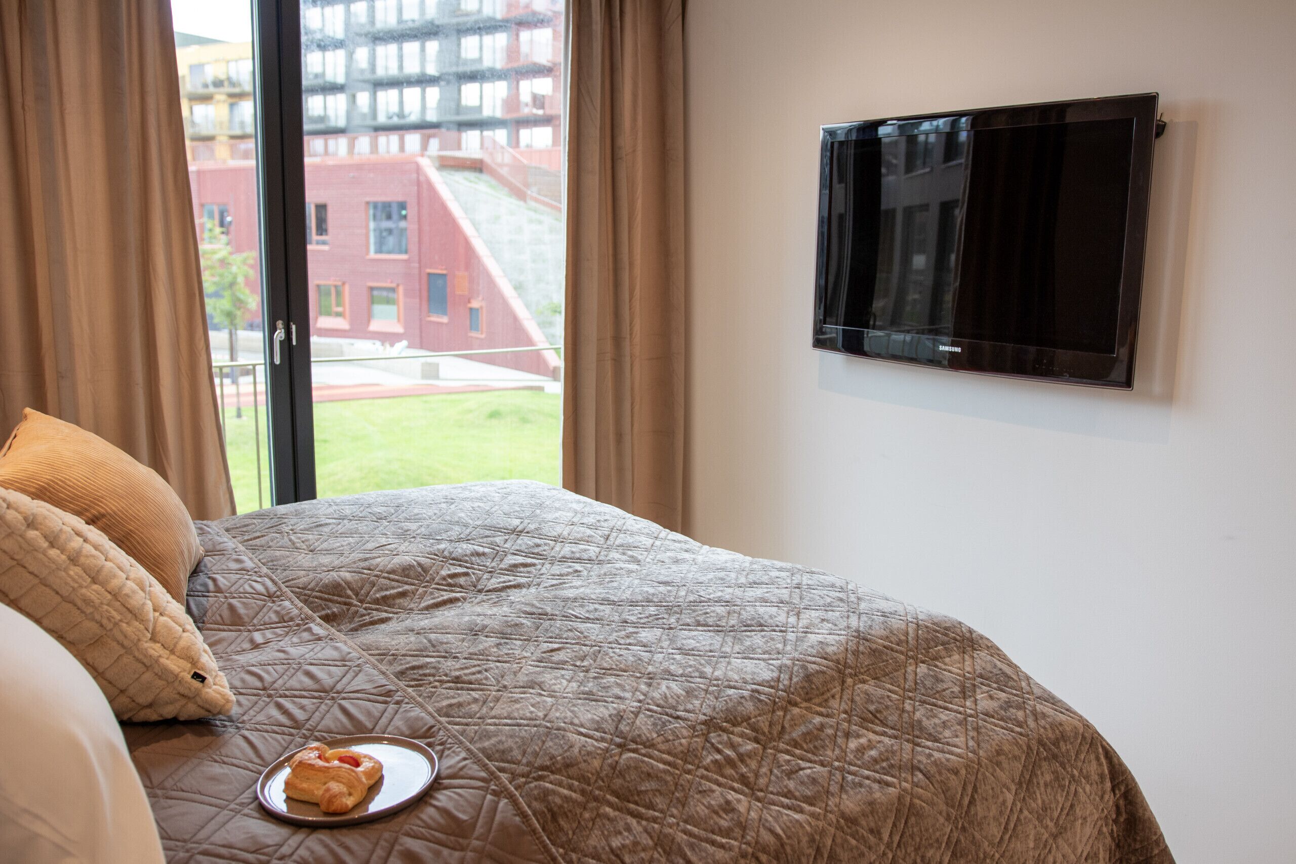 Hotellihuone, jossa on sänky, seinälle asennettu televisio ja ikkuna, josta on näkymä rakennuksiin.