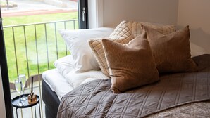 Ein Bett mit gesteppter Decke und mehreren Kissen, ein kleiner Tisch mit Gläsern und einer Flasche sowie Blick auf ein Gebäude durch eine Balkontür.