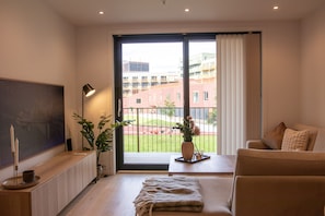 Un soggiorno moderno con un divano, un tavolino, una televisione e una grande finestra con vista su edifici e un giardino.