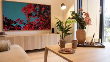 A modern living room with a wooden sofa, a coffee table with pastries, a large floral painting, a potted plant, and a vase with flowers.