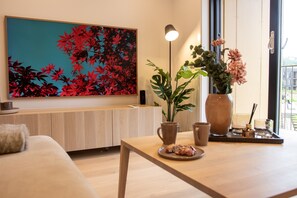 A modern living room with a wooden sofa, a coffee table with pastries, a large floral painting, a potted plant, and a vase with flowers. - Aurora Arctic Apartment (Tromsø)