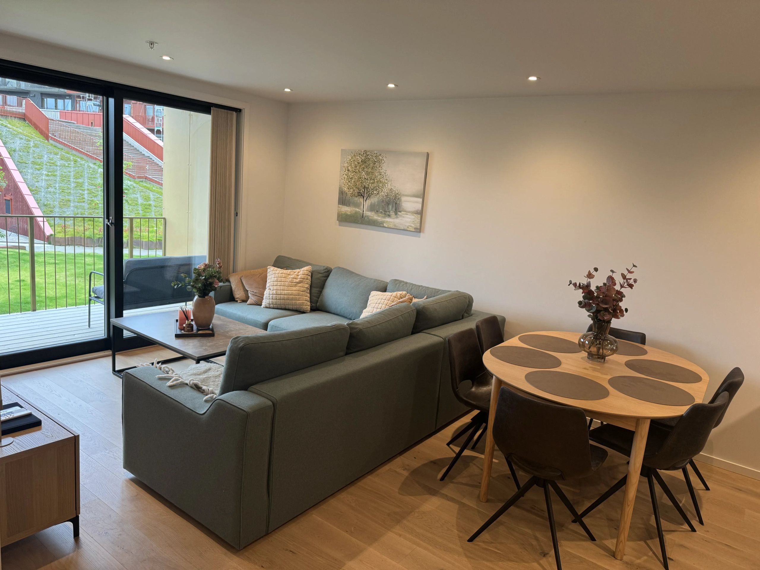A modern living room with a grey sofa, a wooden dining table with chairs, a painting on the wall, and a view of a green outdoor area through a sliding door.