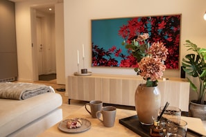 A modern living room with a sofa, a coffee table with a plate of food, two mugs, a vase with flowers, and a painting on the wall. - Aurora Arctic Apartment (Tromsø)