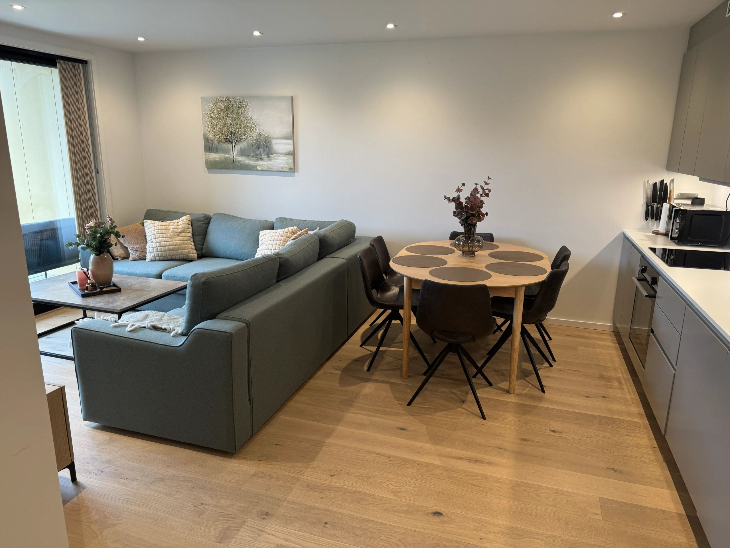 A modern living room with a grey sofa, a round dining table with chairs, and a kitchen area with a microwave.