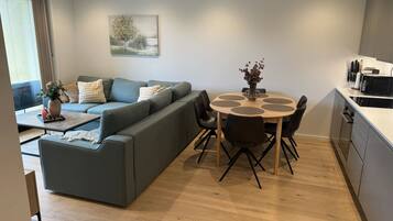 A modern living room with a grey sofa, a round dining table with chairs, and a kitchen area with a microwave.