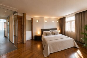 Ein modernes Hotelzimmer mit einem großen Bett, einem Nachttisch mit Lampe, einer Glastür, die auf einen Balkon führt, und Blick auf die Stadt.