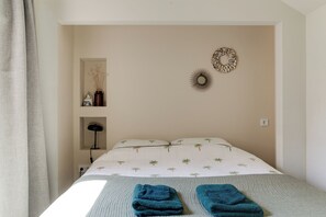 A bedroom with a bed, two wall-mounted decorative items, a shelf with a vase and a lamp, and a window with curtains.
