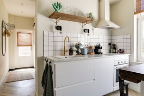A kitchen with white cabinets, a wooden shelf with glassware, a window with blinds, and a door.