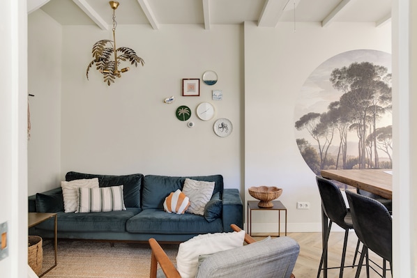 A modern living room with a dark green sofa, a wooden dining table with chairs, a chandelier, and wall art including a large circular painting.