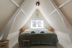 A bedroom with a sloped ceiling, a bed with a green checkered bedspread, two bedside lamps, a small wooden stool, and a window with white blinds.