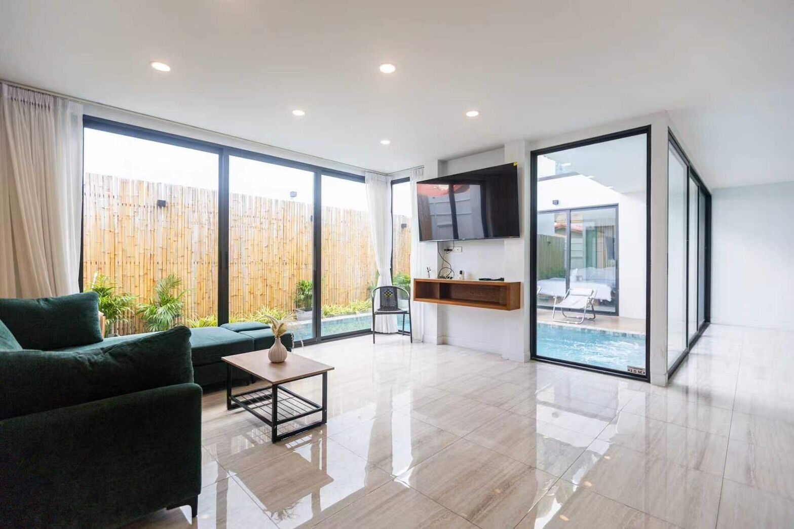 A modern living room with a sofa, a coffee table, a TV mounted on the wall, and a glass door leading to a pool.