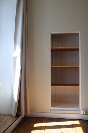 Ein Zimmer mit einer fest installierten Regaleinheit an der Wand, Holzboden und einem Fenster mit Vorhängen.