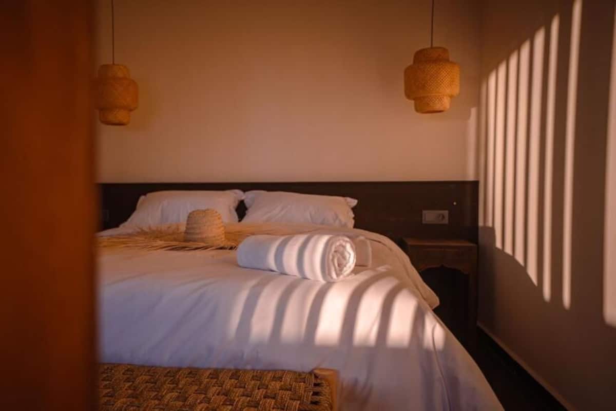 Una cama bien hecha con sábanas blancas, una cesta tejida y dos lámparas fijadas a la pared.
