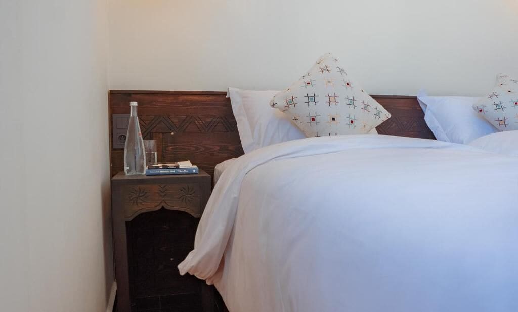 Een net opgemaakt bed met witte lakens en gestreepte kussens, een houten nachtkastje met een glazen fles en boeken, en een eenvoudig wandontwerp.