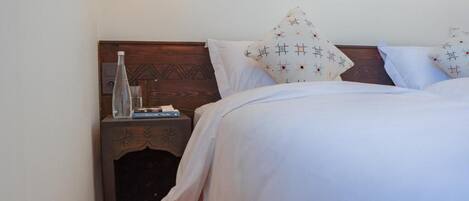 Una cama bien tendida con ropa de cama blanca y almohadas estampadas, una mesita de noche de madera con una botella de vidrio y libros, y un diseño de pared sencillo.