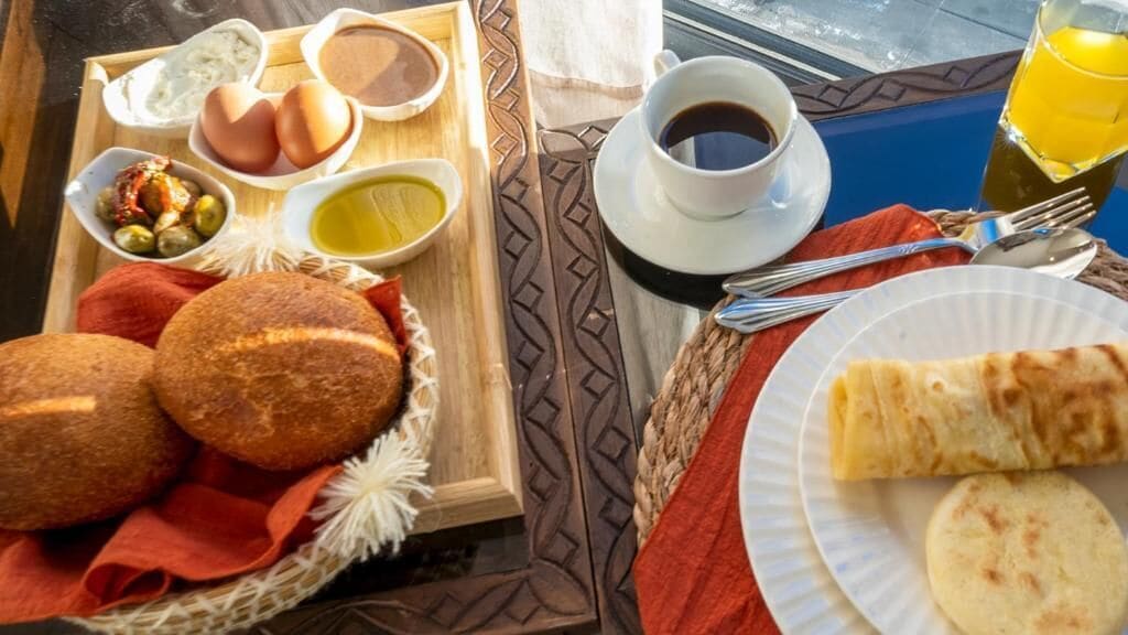 Bandeja de desayuno con pan, huevos, aceitunas y salsas, una taza de café y un vaso de zumo de naranja.