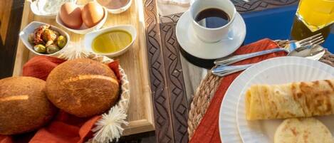Bandeja de desayuno con pan, huevos, aceitunas y salsas, una taza de café y un vaso de zumo de naranja.