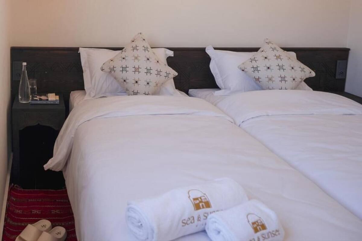 Una cama doble con ropa de cama blanca y almohadas estampadas, una mesita de noche con una botella y un libro, y un par de zapatillas en el suelo.