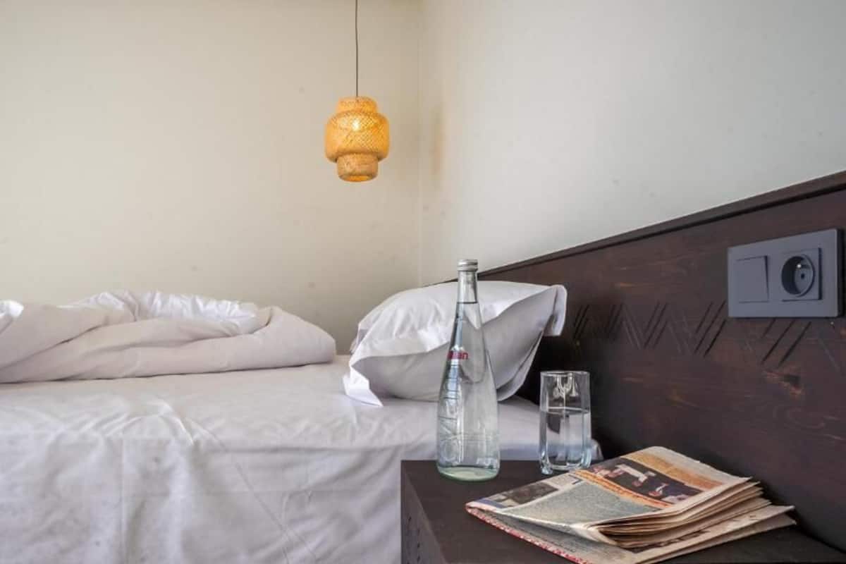 Una cama con sábanas blancas, una botella y un vaso sobre una mesita de noche de madera.