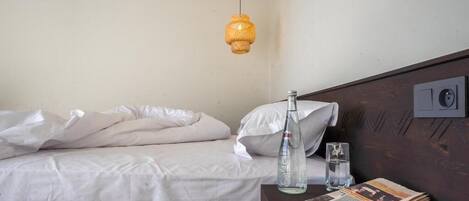 Una cama con sábanas blancas, una botella y un vaso sobre una mesita de noche de madera.